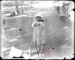 [Little girl with doll's wagon in yard]